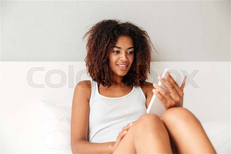 Smiling confident woman in white pajamas chatting on smartphone at home in the morning, stock photo