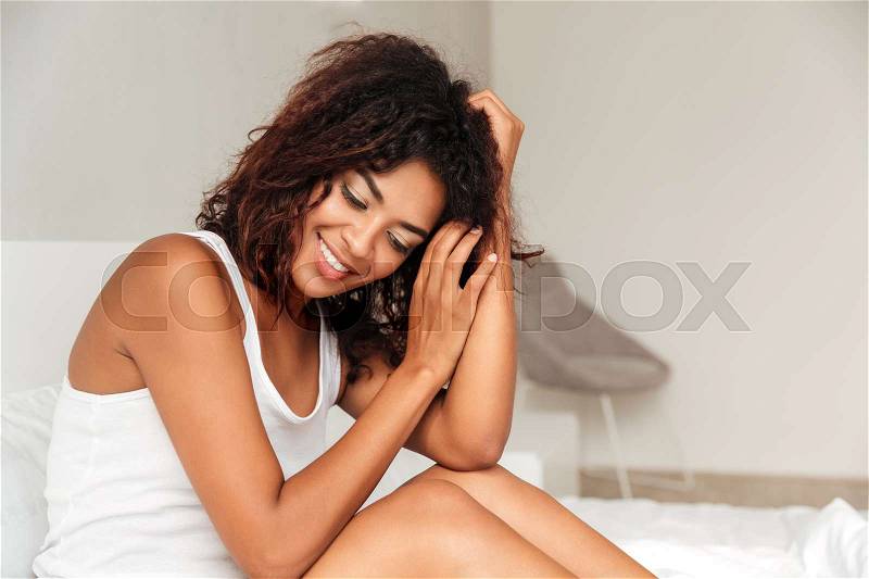Smiling african woman with curly hair and closed eyes sitting in bed in the morning, stock photo
