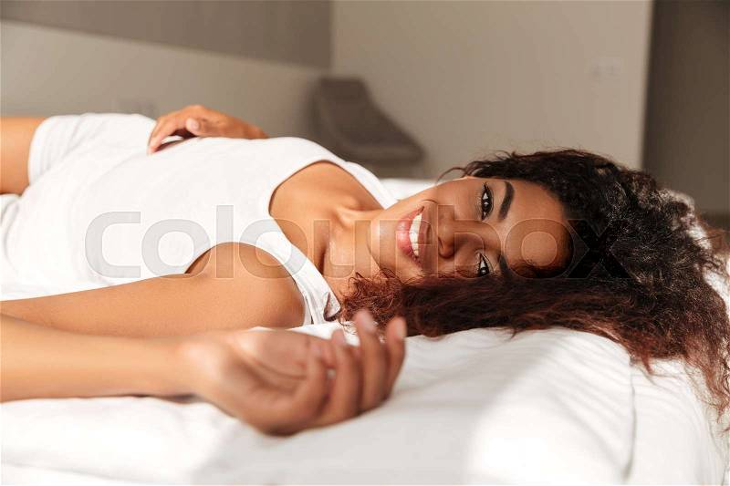 Cheerful sleepy african woman lying in bed in the morning in the sun, stock photo