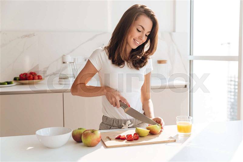 Smiling young woman cutting fruits on a wooden board while making breakfast in a kitchen, stock photo