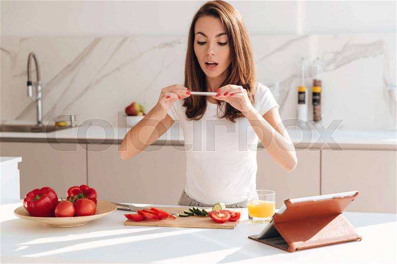 Happy smiling woman taking a picture of vegetable slices on a wooden board with mobile phone while standing at the kitchen, stock photo