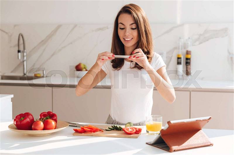 Smiling casual woman taking a picture of sliced vegetables on a wooden board with mobile phone while standing at the kitchen, stock photo