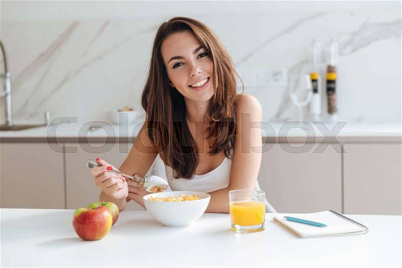 Smiling healthy woman eating corn flakes cereal while sitting and having breakfast at the kitchen table, stock photo