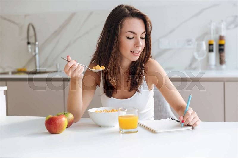 Smiling attractive woman eating corn flakes cereal for breakfast while sitting and writing in notepad at the kitchen table, stock photo