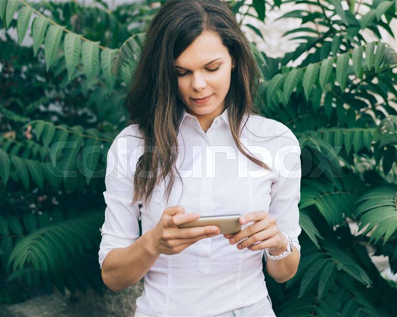Young woman in a white blouse checks her phone against a background of green leaves in the park, stock photo
