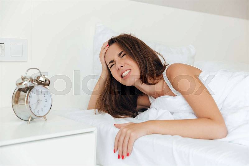 Angry annoyed woman laying in bed in the morning with her ringing alarm clock, stock photo