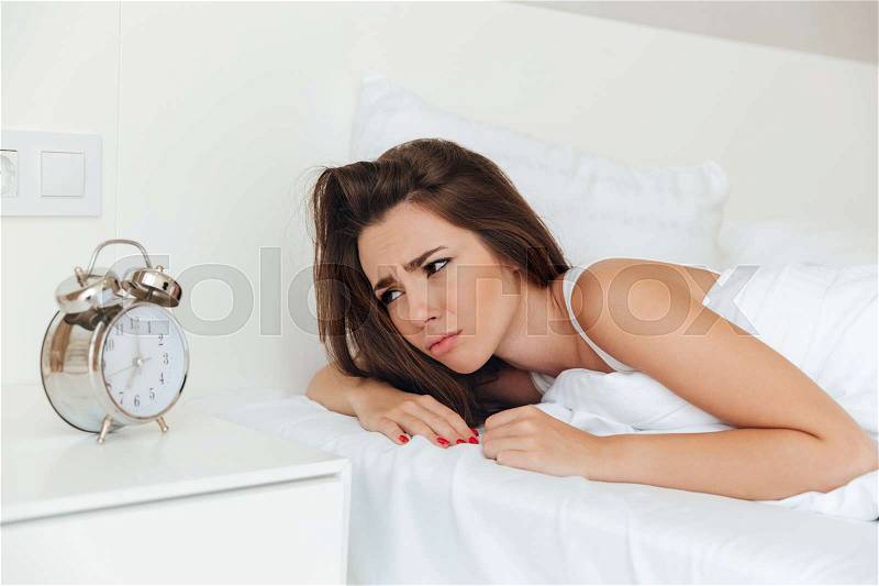 Upset sleepy woman laying in bed in the morning and looking at ringing alarm clock, stock photo
