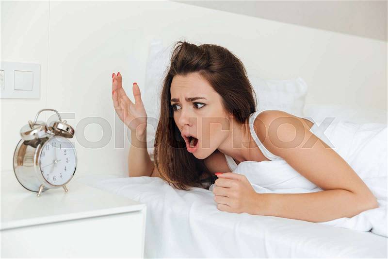 Confused woman laying in bed in the morning and looking at ringing alarm clock, stock photo