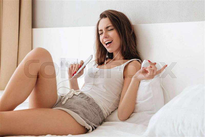 Young pretty woman in earphones listening to music and singing while laying on bed and holding mobile phone, stock photo