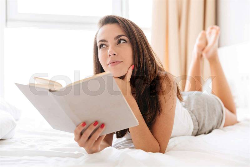 Pensive pretty woman holding a book while laying on bed at home and looking away, stock photo