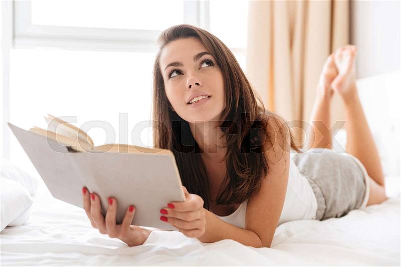 Thoughtful pretty woman holding a book while laying on bed at home and looking away, stock photo