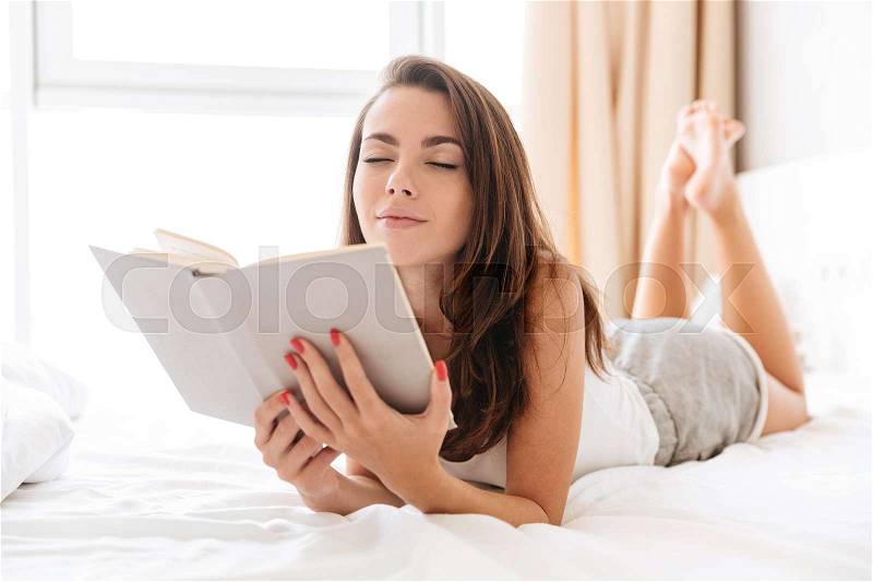 Lovely young woman holding a book while laying on bed at home with eyes closed, stock photo