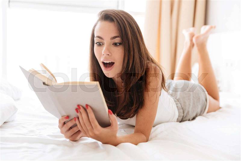 Smiling surprised girl reading book while laying on bed at home, stock photo