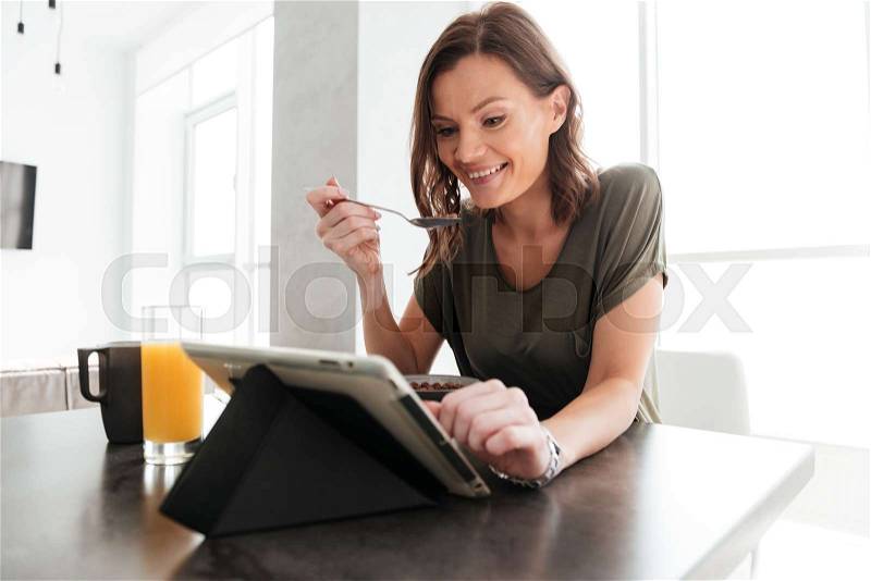 Happy casual woman eating by the table on kitchen and looking at the tablet computer, stock photo
