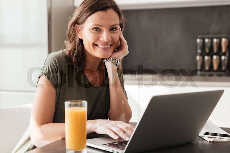 Happy casual woman sitting by the table with juice and tablet computer on kitchen, stock photo