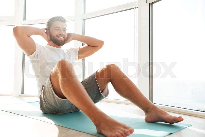 Smiling Handsome bearded man doing abdominal exercises near the window at home, stock photo