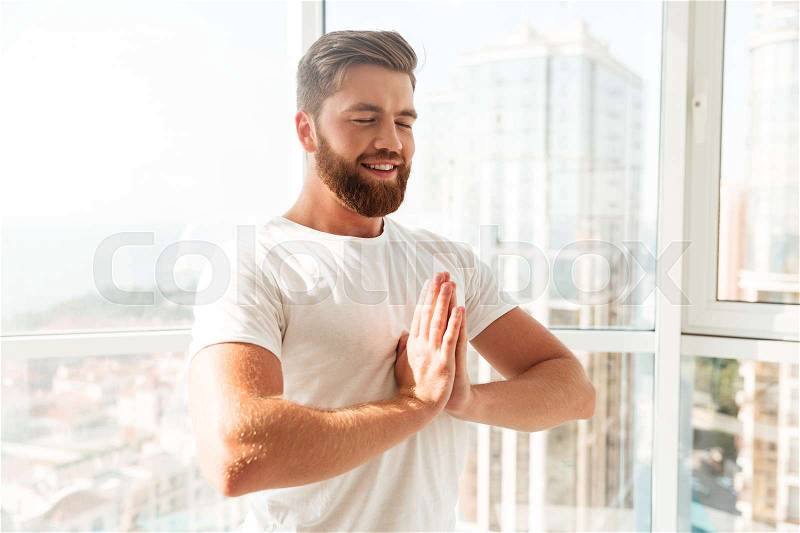 Side view of smiling bearded man meditation with closed eyes over window background at home, stock photo