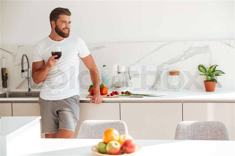 Smiling bearded man standing on kitchen with ice drink and looking away, stock photo
