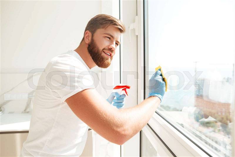 Side view of smiling bearded man wipes a window at home, stock photo
