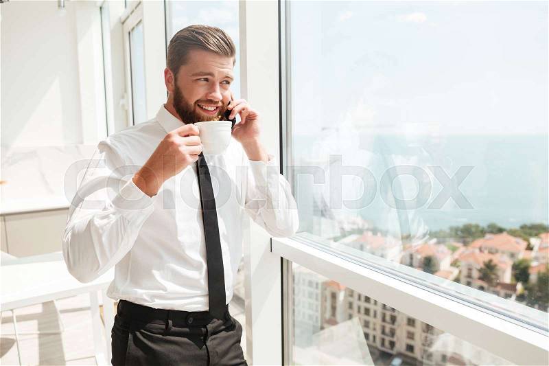 Happy business man drinking coffee and talking by the smartphone near the window, stock photo