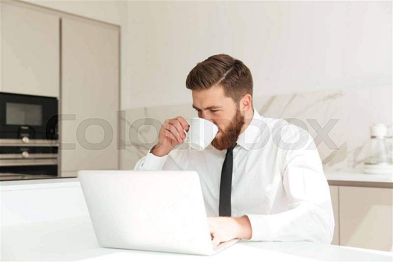 Smiling Bearded business man drinking coffee and using laptop computer by the table in kitchen, stock photo