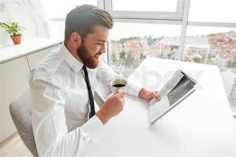 Side view of smiling bearded man reading journal and drinking coffee by the table on kitchen, stock photo