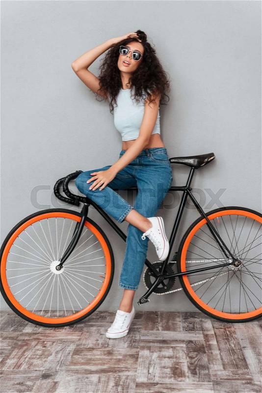 Full length image of beauty curly woman in sunglasses posing with bicycle over gray background, stock photo