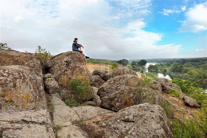 The teenager sits on top of a large stone boulder on the bank of the South Bug River and looks at the river below. The river Southern Bug in the summer - river flow, rocky shores, bright green vegetation and a cloudy blue sky, stock photo
