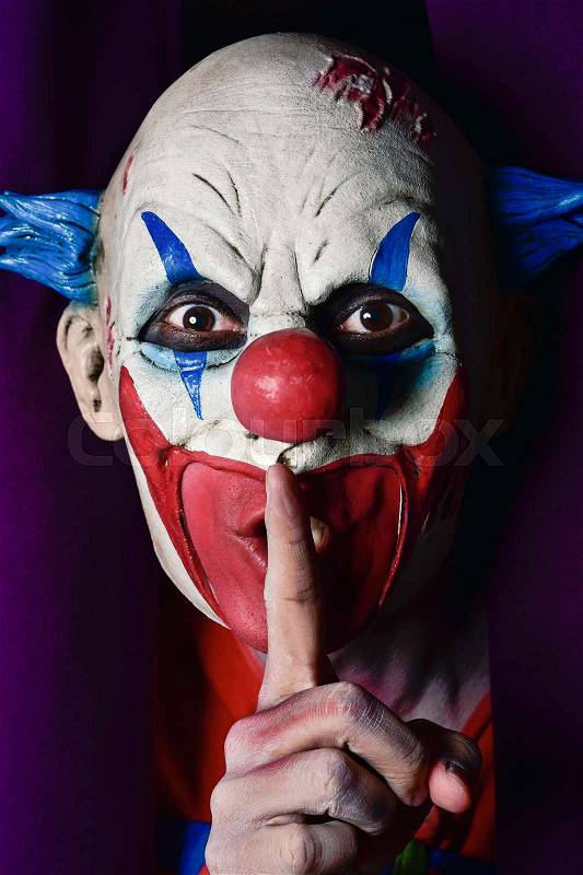 A scary evil clown peering out from a purple stage curtain, with his forefinger in front of his lips, asking for silence, stock photo