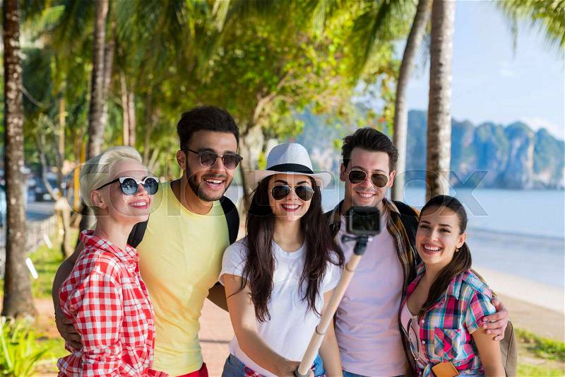 People Group Take Selfie With Action Camera On Stick While Walking In Palm Tree Park On Beach, Happy Smiling Mix Race Friends On Summer Vacation, stock photo