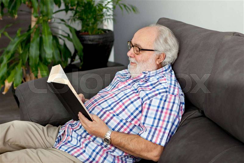 Retired man reading a book on the sofa in his home, stock photo