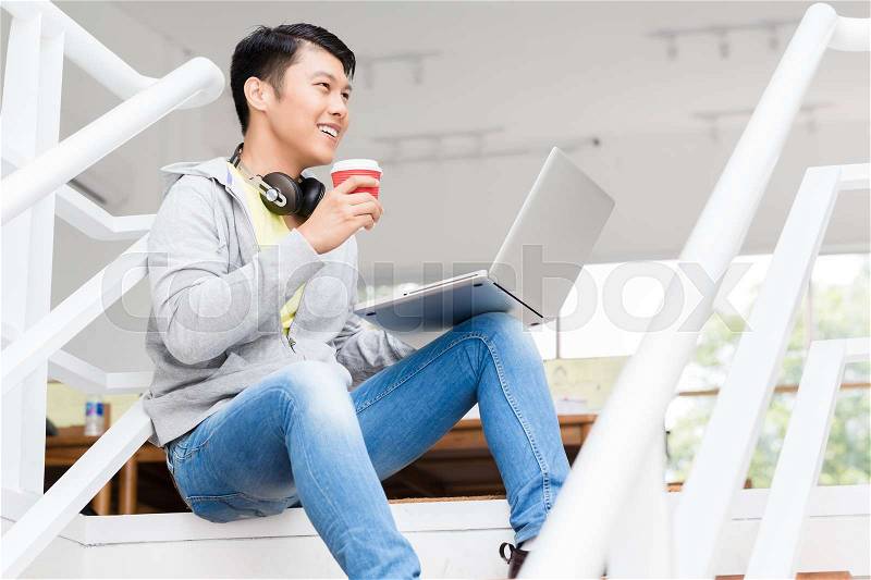 Happy young Asian employee drinking coffee while using a laptop in the interior of a modern office, stock photo