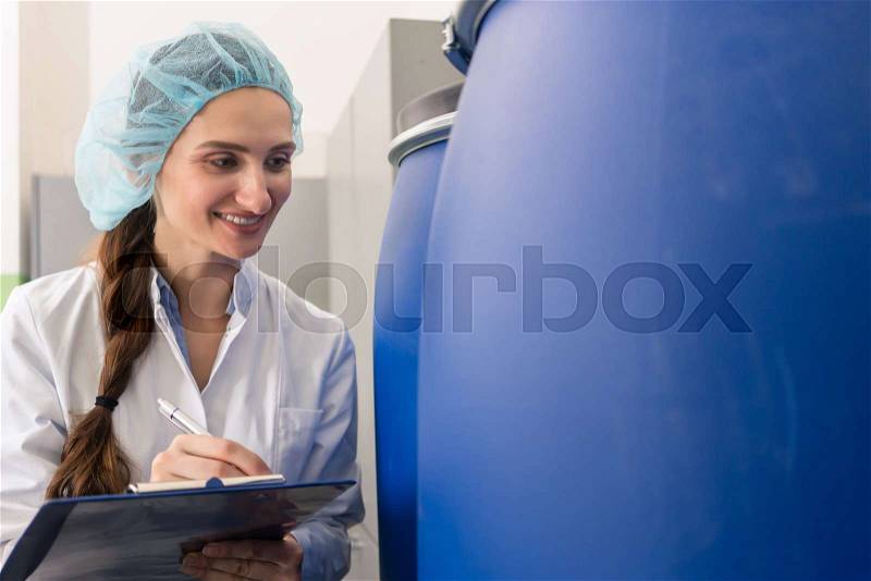 Motivated manufacturing employee writing a report while checking the plastic containers, with liquid cosmetics during quality control in the factory, stock photo