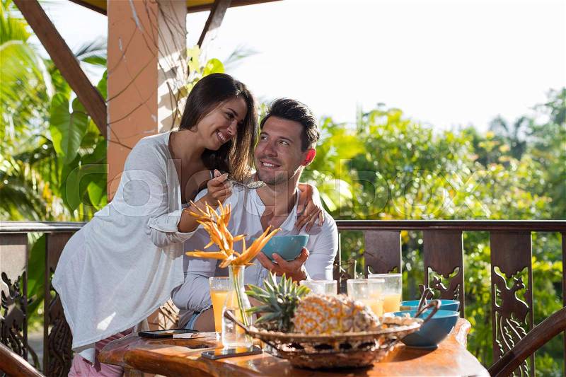 Couple On Summer Terrace Having Breakfast, Woman Feeding Man With Porridge In Morning Eating Outdoors With View On Tropical Forest, stock photo