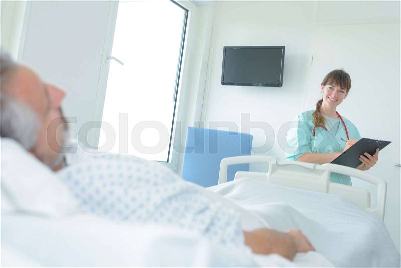 Smiling nurse comforting senior man at bed in hospital, stock photo