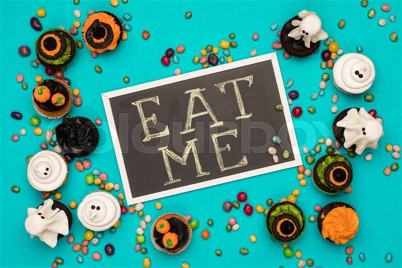 Top view of various decorative halloween cupcakes, eat me inscription and colorful candies on blue, stock photo