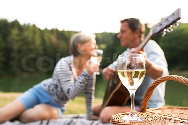 Beautiful senior couple at the lake having a picnic, sitting on blanket, drinking wine. Man playing guitar, stock photo