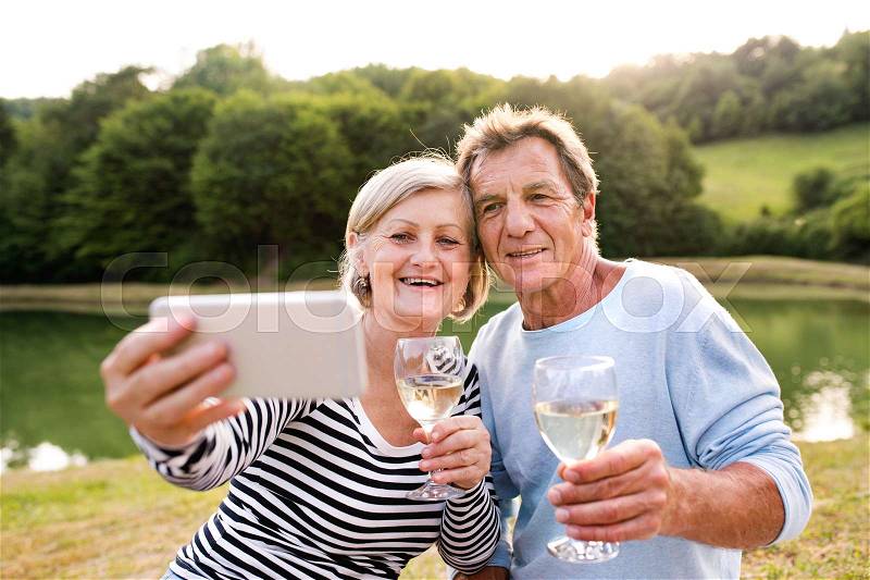 Beautiful senior couple at the lake having a picnic, drinking wine, holding smart phone, taking selfie, stock photo