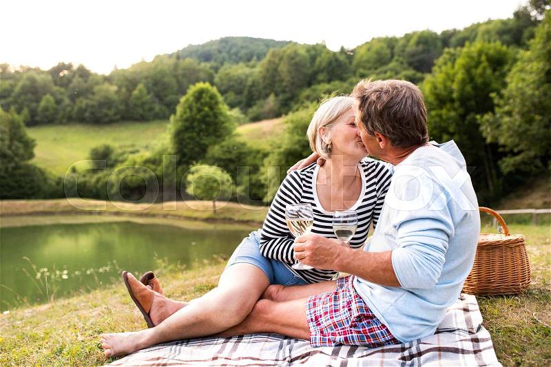 Beautiful senior couple at the lake having a picnic, sitting on blanket, drinking wine and kissing, stock photo