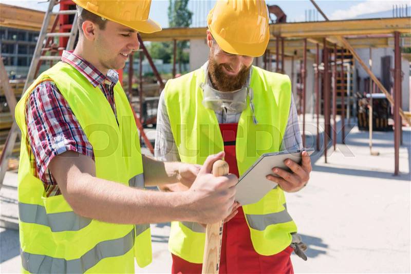 Two young construction workers smiling while using a tablet for online communication through social media during break at work, stock photo