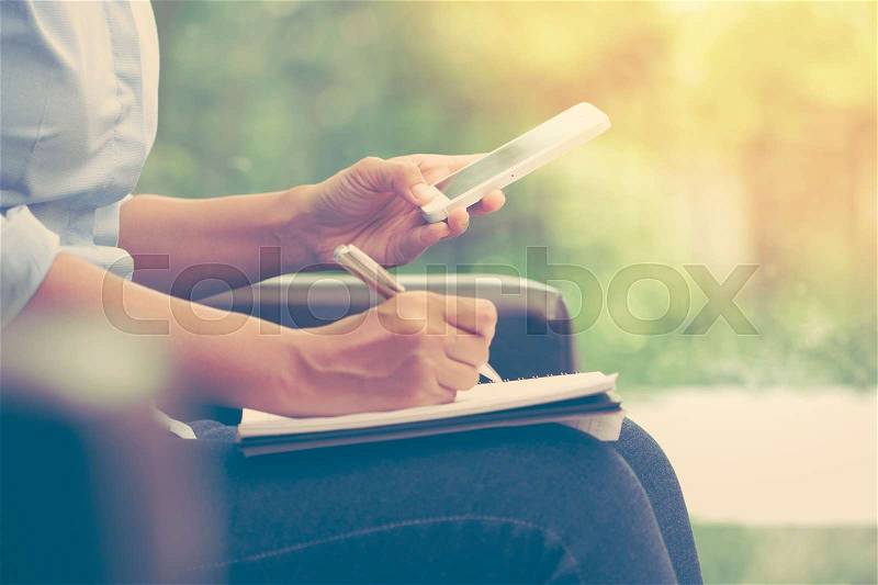 Business woman using mobile smart phone and writing on notebook at home office, stock photo
