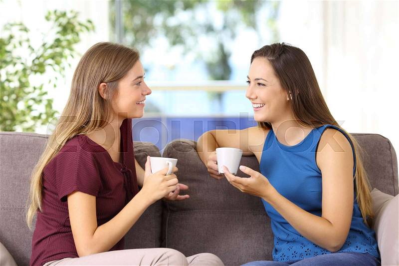 Two happy girls talking and drinking sitting on a sofa at home, stock photo
