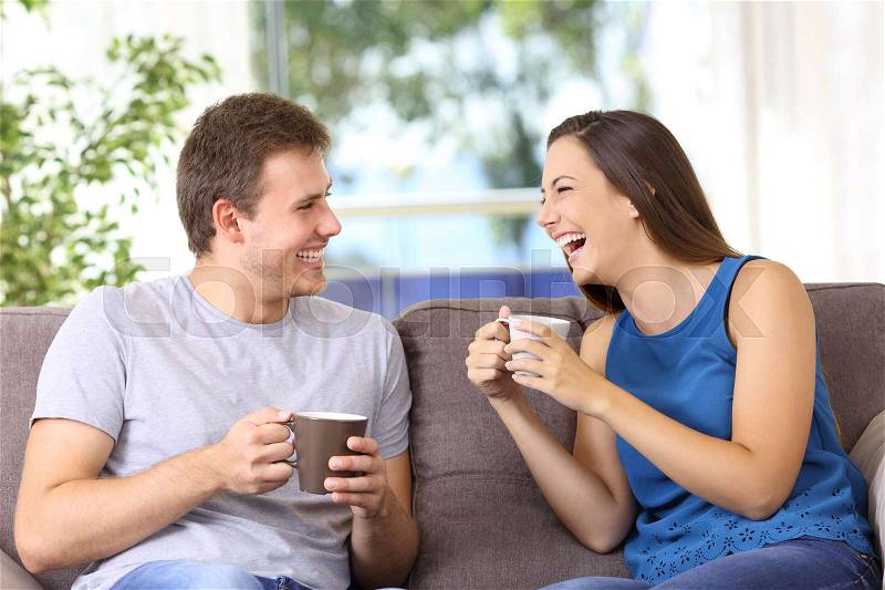 Two people talking and laughing loud sitting on a sofa at home, stock photo