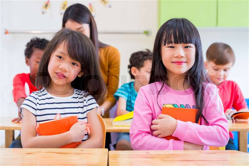 Two Asian girl kid hugging book and smiling in classroom and while teacher teach friends beside them,kindergarten education, stock photo