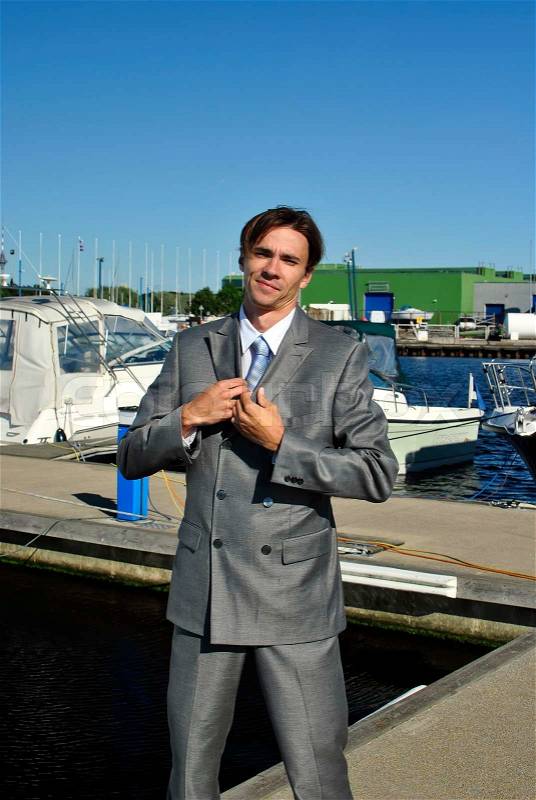 Man in a gray business suit straightens his tie, in the background of yachts, stock photo