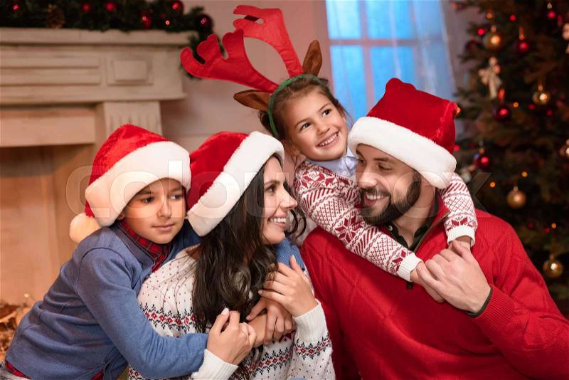 Cheerful young family in santa hats and antlers smiling each other and hugging at home, stock photo