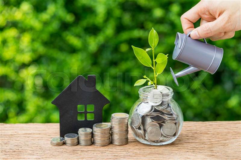 Stack of coins, paper house and growth sprout plant with hand watering as business finance or grow investment concept, stock photo