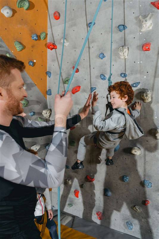 Little boy in harness hanging on a climbing wall and high-fiving father, stock photo