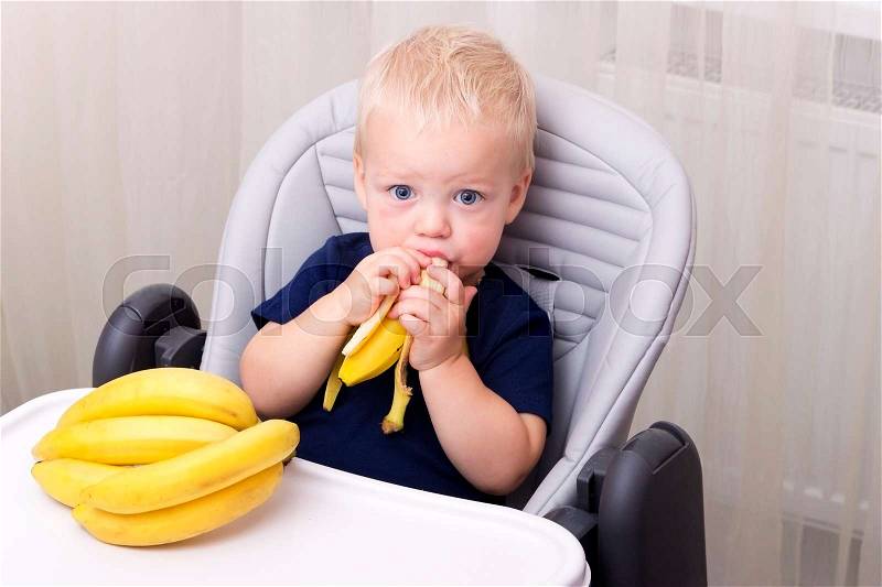 Cute one year old toddler eating a banana and sitting in the baby chair, stock photo