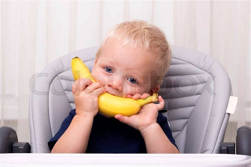 Cute one year old toddler eating a banana and sitting in the baby chair. Funny kid with banana smile, stock photo